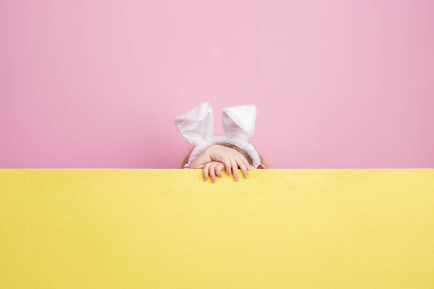Funny little girl with bunny ears on her head hides behind the yellow board against a pink wall - Zdjęcie, obraz