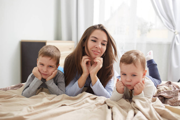 Happy young mother dressed in light blue pajama lays with her two little sons holding their hands under their chins on the bed with beige blanket in the bedroom with big window - Fotoğraf, Görsel