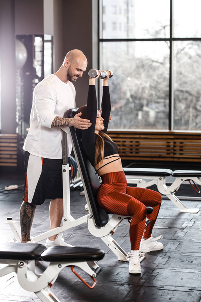 Slender girl in stylish sports clothes sits on a sports bench holding dumbbell in her hands and athletic man stands behind her in the modern gym - Φωτογραφία, εικόνα