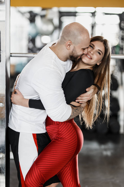 Romantic happy athletic couple. Strong man and slender beautiful girl are hugging in the modern gym next to the sport equipment - Fotografie, Obrázek