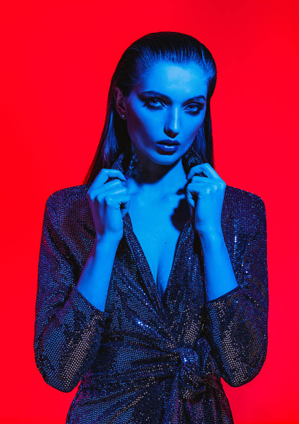 Fashion girl with long hair and stylish makeup in a black shining dress poses on the red background in neon light in the studio - Fotografie, Obrázek