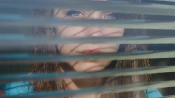 Όμορφη νεαρή γυναίκα κοιτάζει έξω από το παράθυρο μέσα από τις περσίδες - Πλάνα, βίντεο