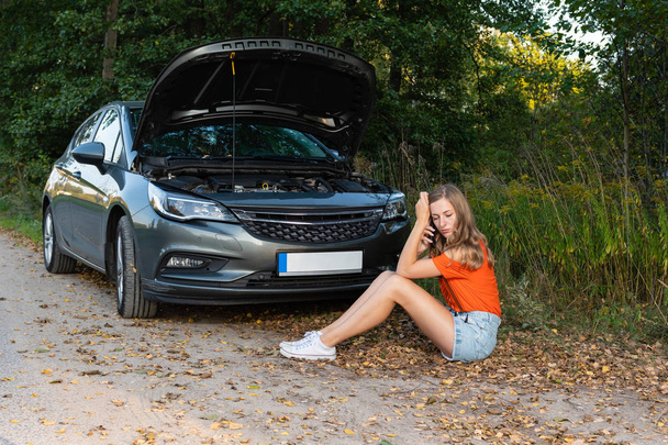 jonge vrouw in de buurt van gebroken auto spreken via de telefoon heeft hulp nodig - Foto, afbeelding