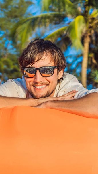 Summer lifestyle portrait of man sitting on the orange inflatable sofa on the beach of tropical island. Relaxing and enjoying life on air bed VERTICAL FORMAT for Instagram mobile story or stories size - Foto, immagini