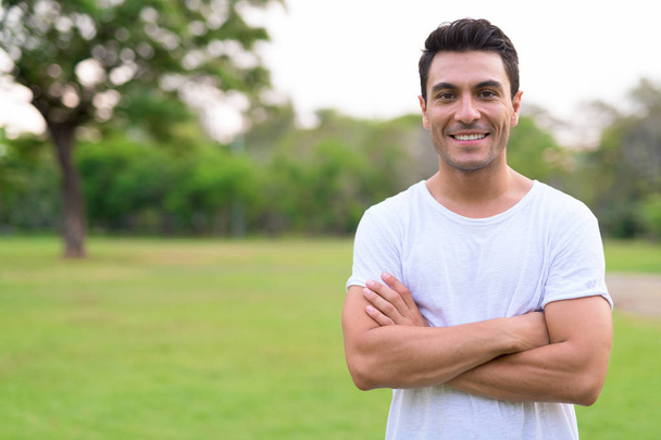 Feliz jovem hispânico sorrindo com os braços cruzados no parque ao ar livre
 - Foto, Imagem