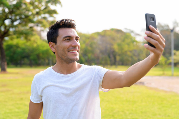 Szczęśliwy młody przystojny mężczyzna Hiszpanie biorąc selfie w parku - Zdjęcie, obraz