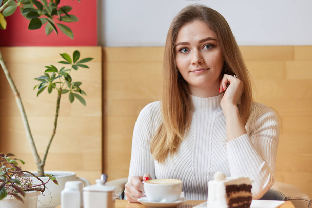 Horizontal shot of beautiful young girl enjoys hot espresso with delicious piece of cake, has pleasant conversation with friend, looks directly at camera, poses over cosy cafe interior, has breakfast - Foto, Imagen