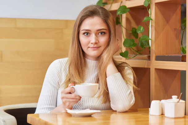 Photo of beautiful young female of European appearance, dressed in white clothes, drinks aromatic hot coffee or tea in cosy cafeteria, looks directly at camera, has good rest during weekend. - Foto, immagini
