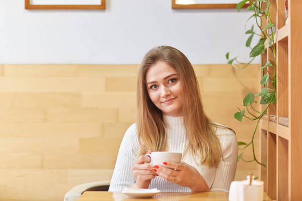Image of lovely young female model with straight light hair, holds mug of drink, has free time, meets with partner in coffee shop, enjoys calm atmosphere, likes drinking coffee. Time for having lunch - Foto, Imagem