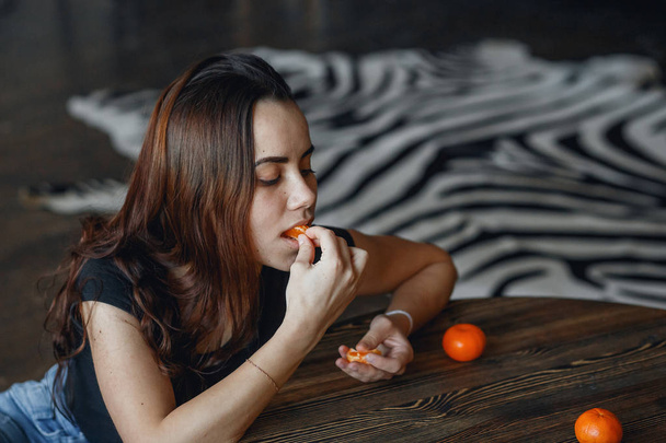 Νεαρό κορίτσι τρώει γλυκά μανταρίνια κάθεται στο πάτωμα κοντά στο τραπέζι - Φωτογραφία, εικόνα