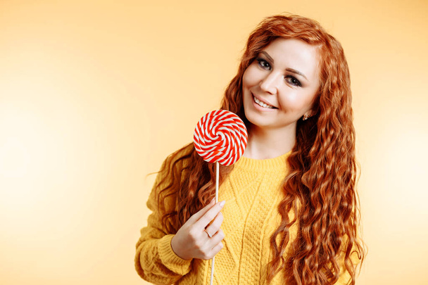 Beautiful young curly redhead woman holding a red white lollipop and smiling - 写真・画像