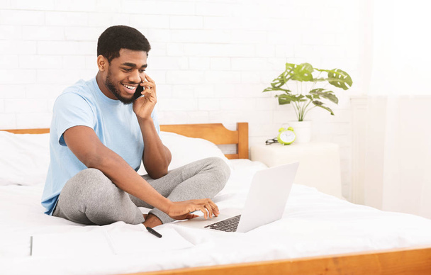 Sonriente hombre hablando por teléfono y utilizando el ordenador portátil en la cama
 - Foto, Imagen