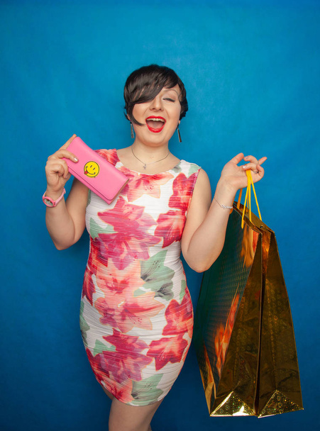 Chica adicta a las compras divirtiéndose comprando todo. se para sobre un fondo azul con un bolso rosa en las manos y bolsas de las tiendas
. - Foto, Imagen