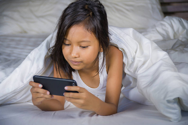 happy and beautiful 7 years old child having fun playing internet game with mobile phone lying on bed cheerful and excited in young girl and technology concept - 写真・画像