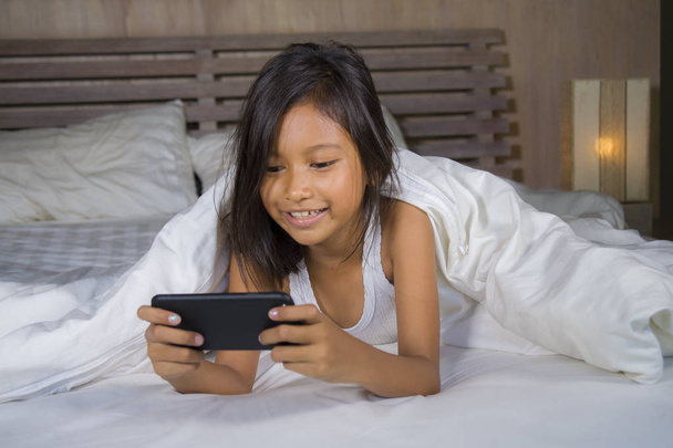 happy and beautiful 7 years old child having fun playing internet game with mobile phone lying on bed cheerful and excited in young girl and technology concept - 写真・画像