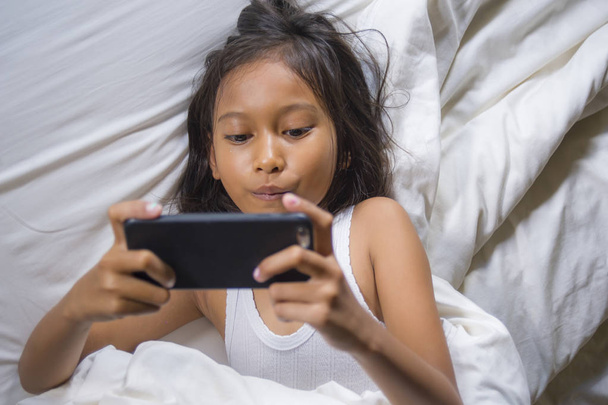 happy and beautiful 7 years old child having fun playing internet game with mobile phone lying on bed cheerful and excited in young girl and technology concept - Photo, Image