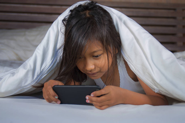 happy and beautiful 7 years old child having fun playing internet game with mobile phone lying on bed cheerful and excited in kid and domestic technology concept - 写真・画像