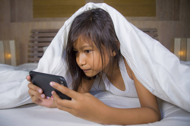 happy and beautiful 7 years old child having fun playing internet game with mobile phone lying on bed cheerful and excited in kid and domestic technology concept - 写真・画像