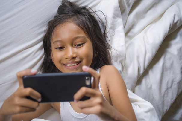 happy and beautiful 7 years old child having fun playing internet game with mobile phone lying on bed cheerful and excited in kid and domestic technology concept - Foto, imagen