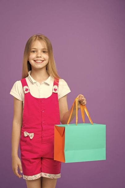 Little child with shopping bag on violet background. Happy girl smile with paper bag. Kid shopper in fashion jumpsuit. Holidays and celebration. Satisfied with her shopping. sale and black friday - Foto, Imagem