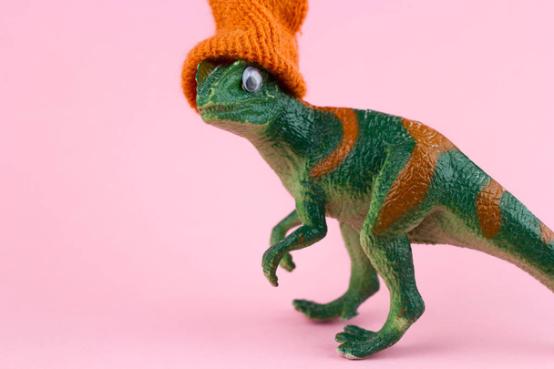funny green dinosaur toy in little knitted orange hat  near on pastel pink background - Foto, Imagen