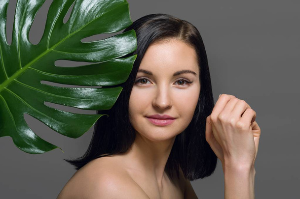Studio beauty portrait of young brunette with natural make-up perfect skin with green exotic leaf on gray background, copy space. Concept of natural cosmetics, face and body care products. - Photo, image