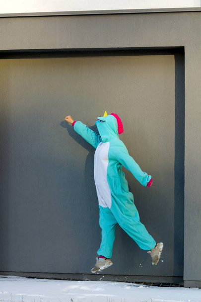 cute woman in kigurumi unicorn costume jump like superhero against gray wall on the street - Photo, Image