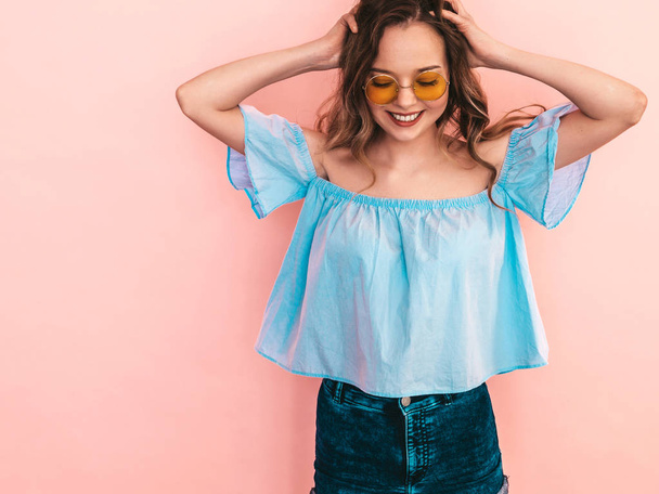 Portrait of beautiful smiling cute model in round sunglasses. Girl in summer hipster colorful clothes. Model posing on pink  background. Playing with her hair - 写真・画像