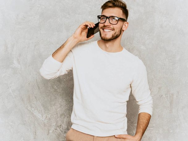 ハンサムな笑顔ヒップスター lumbersexual ビジネスマン モデル カジュアルな夏の白い服を着ての肖像画。ファッション スタイリッシュな男の灰色の壁に対してポーズします。眼鏡で携帯電話で話す - 写真・画像
