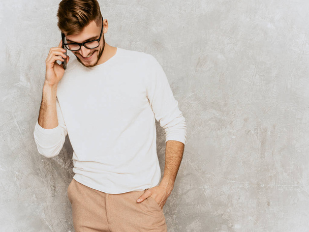 Portrait of handsome smiling hipster lumbersexual businessman model wearing casual summer white clothes. Fashion stylish man posing against gray wall. Speaking on mobile phone in spectacles - Foto, imagen