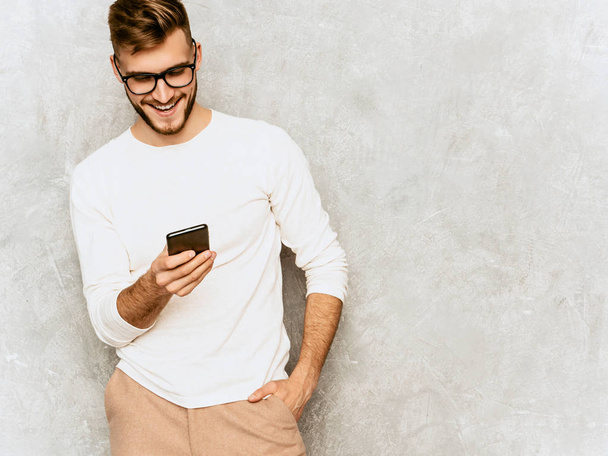 Portrait of handsome smiling hipster lumbersexual businessman model wearing casual summer white clothes. Fashion stylish man posing against gray wall. With mobile phone in spectacles - Zdjęcie, obraz