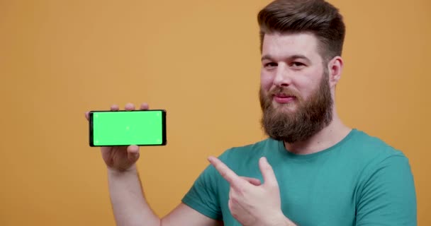 Joven barbudo apuntando con el dedo a su teléfono inteligente con pantalla verde en
 - Metraje, vídeo