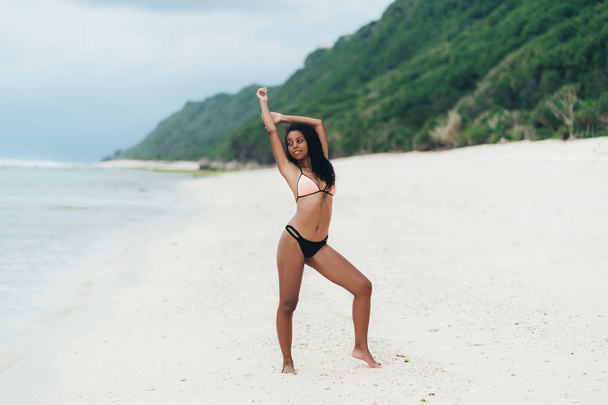 Slender sexy dark skinned girl in swimwear posing on beach with sand. Afro american woman resting on paradise island - Фото, зображення