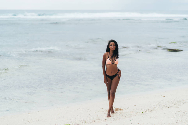 Sexy african american girl in swimwear resting on ocean beach. Young black skinned woman with curly hair stands on seashore - Photo, Image
