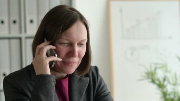 Veselá podnikatelka mluvit na mobilním telefonu při sezení u stolu v obchodní kanceláři, zpomalené záběry z digitální - Záběry, video