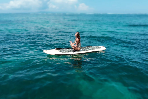若い美しい女性のスーペリアー paddleboarding で海で瞑想します。健康的なライフ スタイル。穏やかな水でリラックスできるヨガのポーズの女の子 - 写真・画像