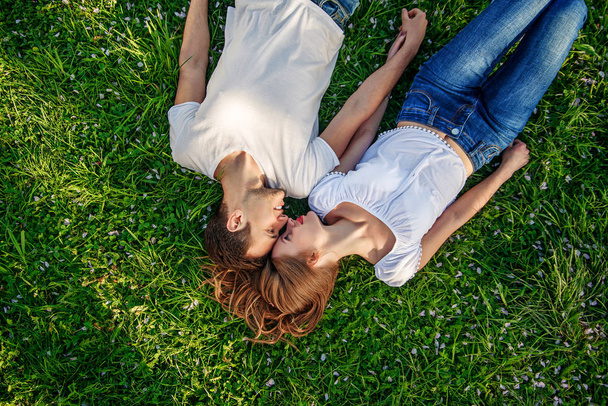 Romantic couple of young people lying on grass in park. They lay on the shoulders of each other and hold hands together. They look happy. View from above. - Foto, Imagen