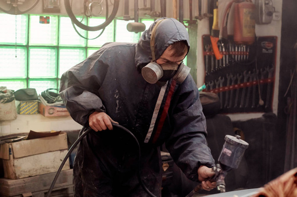 Mann mit Schutzkleidung und Maske malte Auto mit Sprühkompressor. - Foto, Bild
