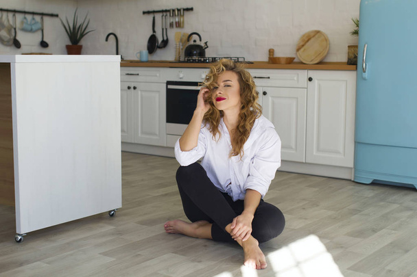 Portrait of young blonde woman at bright kitchen sitting on the floor - Photo, image