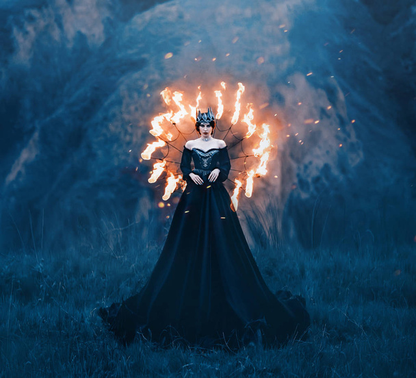 dark queen of night, majestic priestess in long black dress with bare shoulders, witch raven in a burning spider web, powerful devil with metal cold crown on her head, mistress and fire goddess - Foto, imagen