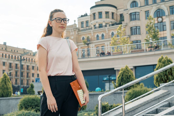Outdoor portrait of female student 16, 17 years old. Girl in glasses, with backpack, textbooks. City background - Fotó, kép