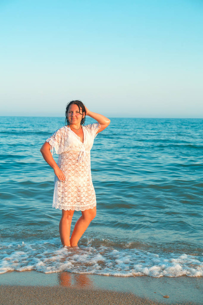 Egy fiatal nő ül a homok, a tenger a fehér csipke köpeny a parti hullámok. - Fotó, kép