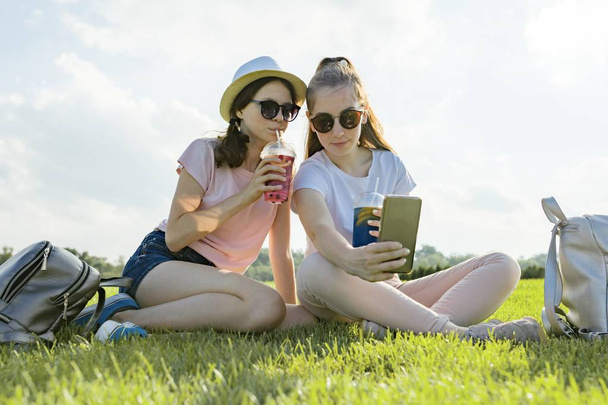Teens girls in hat and sunglasses on the green lawn in the recreation park enjoying their cocktails, golden hour - 写真・画像