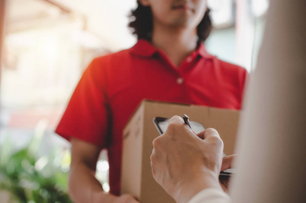 young woman customer appending signature in digital mobile phone receiving parcel post box from courier with home delivery service man in red uniform at home, express delivery, online shopping concept - 写真・画像