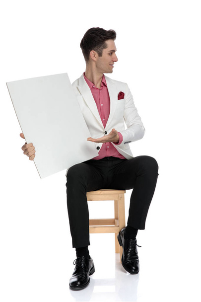 joven hombre de negocios sentado y presentando un tablero en blanco al lado
 - Foto, Imagen
