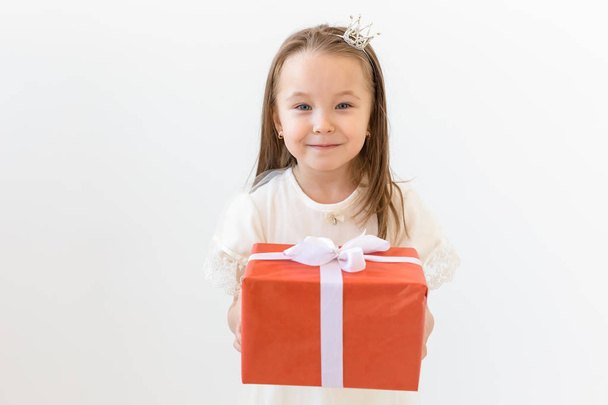 Menschen, Kinder und Urlaubskonzept - Porträt eines glücklichen kleinen Mädchens, das eine Geschenkschachtel über weißem Hintergrund hält - Foto, Bild