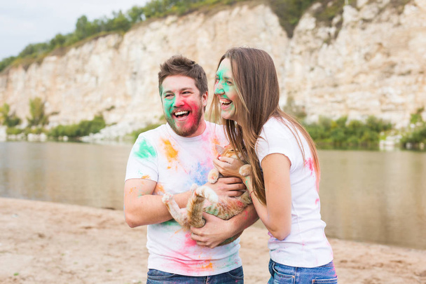 Festival holi, holidays, tourism and nature concept - Couple dressed in white shirts holding cat and covered with colorful dust - 写真・画像