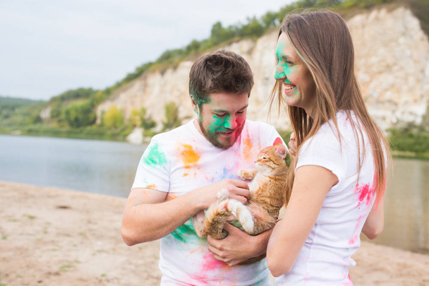 Festival holi, holidays, tourism, pet and nature concept - Portrait of woman and man with cat covered multi-colored dust - Photo, Image