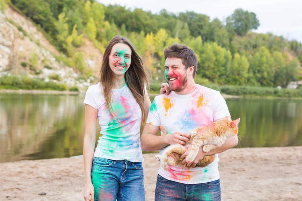 Festival holi, holidays, tourism, pet and nature concept - Portrait of woman and man with cat covered multi-colored dust - Фото, зображення