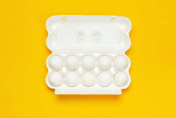 Белые куриные яйца в белой упаковке на желтом фоне сверху плоского вида лежат в копировальном пространстве. Яйца в коробке, натуральная здоровая пища и органическое земледелие. Креативные продукты питания минималистский фон, Пасха
 - Фото, изображение